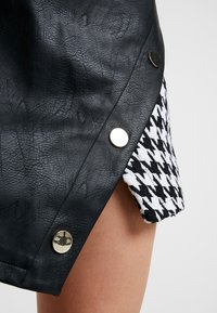 Jupe en simili cuir noire ornée de quincaillerie en argent, avec un tissu à motif pied-de-poule qui apparaît sur le côté. Design de courte longueur.