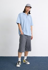 Ljusblå pikétröja, grå denimshorts och mångfärgade sneakers. Tillbehör inkluderar en grårandig keps. Modellen står mot en neutral bakgrund.