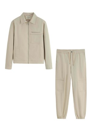 Veste beige légère avec un col, fermeture éclair à l'avant et poche poitrine, assortie à un jogging à taille élastique avec poches latérales.