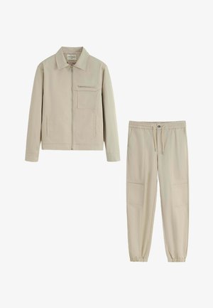 Veste beige légère avec un col, fermeture éclair à l'avant et poche poitrine, assortie à un jogging à taille élastique avec poches latérales.