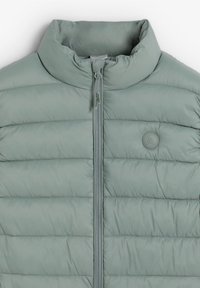 Vaahtomuovilla vuorattu takki vaaleanvihreä, jossa on korkea kaulus, etuvetoketju ja pyöreä logo-ominaisuus. Sileä pinta, jossa on vaakasuoraa ompeleita.