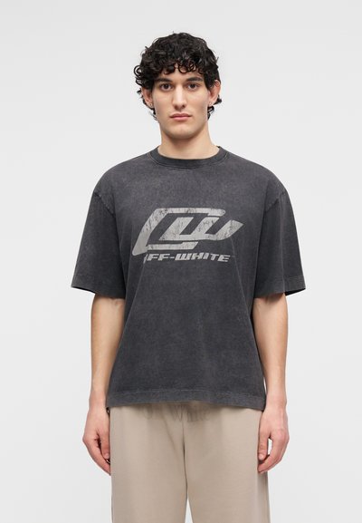 Νέος άνδρας με σκούρα σγουρά μαλλιά που φοράει μια ξεθωριασμένη μαύρη μπλούζα με το λογότυπο Off-White και ανοιχτό μπεζ παντελόνι, στέκεται μπροστά σε απλό φόντο.