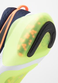 Sportovní bota s tmavě modrým síťovinovým svrškem, jasně oranžovými akcenty, průhlednou neonově zelenou podrážkou a strukturovaným černým zadním panelem.