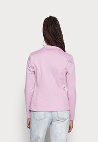 Lila sovittelu takki, jossa on lovettu kaulus, rakenteelliset saumat, pitkät hihat ja sileä pinta, yhdistettynä vaaleisiin farkkuihin.