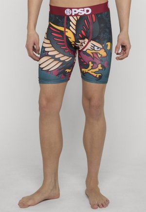 Slank mann iført flerfargede boxer briefs med stort ørnedesign og PSD-logo på burgunder midjebånd, stående barbeint.
