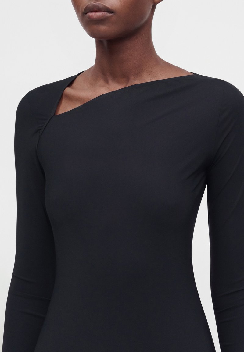 Femme portant un haut noir ajusté à manches longues avec un décolleté asymétrique laissant apparaître une partie de l'épaule et de la clavicule gauche.