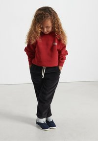 Raudonas džemperis su "America Vintage" logotipu, juodi aksominiai keliaraiščiai su raišteliu, dryžuotos kojinės ir tamsiai mėlyni slip-on batai.