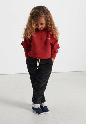 Červená mikina s logom "America Vintage", čierne nohavice z korduroy s šnúrkou, pruhované ponožky a námornícke slip-on topánky.