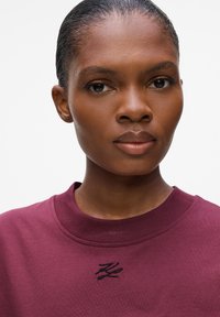 Burgunder Sweatshirt mit rundem Halsausschnitt und schwarzem gesticktem Logo auf der Brust. Glatte Stofftextur, taillierte Passform.