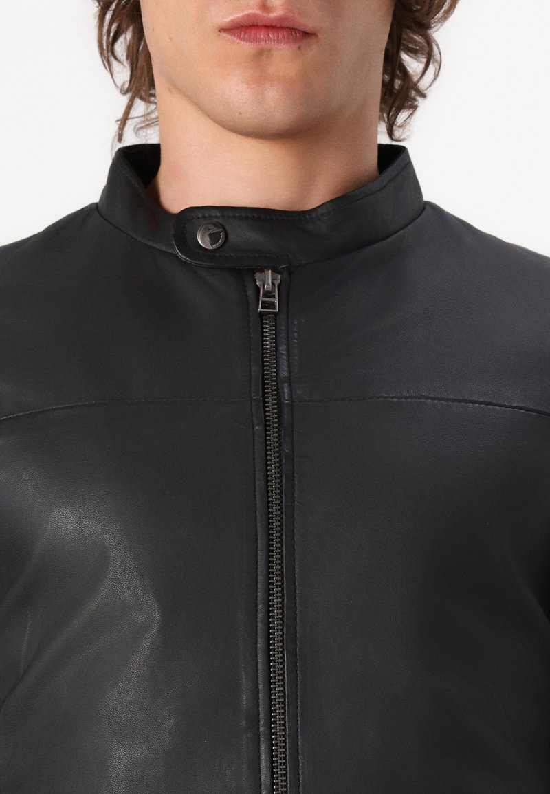 Schwarze Lederjacke mit hohem Kragen, Frontreißverschluss und Druckknopfverschluss. Glatte Textur mit minimalen Nähdetails.