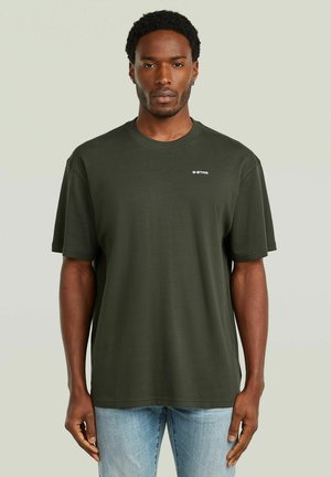 Man draagt een donkergroen G-Star t-shirt en lichtblauwe spijkerbroek, staand tegen een effen lichte achtergrond.