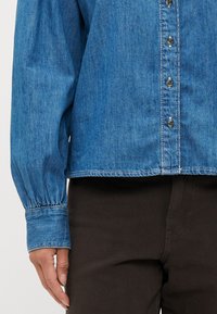 Denimskjorte med knapplukking foran, lange ballongsleeves og sammenkrøllede mansjetter. Blå farge med synlig sømdetalj.