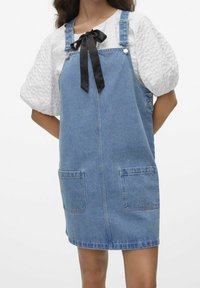 Kvinna som bär en ljusblå denimpinafore-klänning över en vit, texturerad blus med puffärmar, svart snodd knuten vid halsen, händerna bakom ryggen.