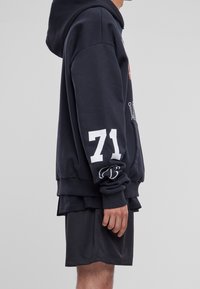 Hombre con sudadera negra con el número blanco "71" en la manga y pantalones cortos de malla negros, de pie de lado contra un fondo gris neutro.