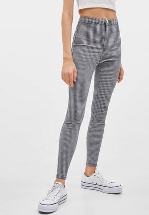 Vrouw draagt een zwart-wit geruite skinny broek met hoge taille, witte platform sneakers en een cropped wit topje, staand tegen een witte achtergrond.