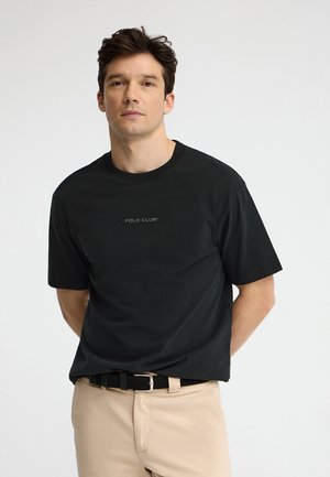 Homme portant un t-shirt noir "Polo Club" et un pantalon beige, debout les mains dans le dos sur un fond blanc uni.