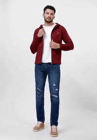 Red quilted zip-up hoodie with kangaroo pockets over a white t-shirt, paired with distressed blue jeans and tan sneakers.