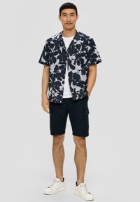 Koszula z krótkim rękawem w granatowy wzór kwiatowy z białym nadrukiem, połączona z ciemnymi krótkimi spodniami typu cargo i białymi sneakersami, o luźnym kroju i detalach kieszeni.