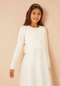 Fehér kardigán hosszú ujjakkal, formázott dizájnnal és díszítő varrással a vállaknál, egy átlátszó gombbal zárva.