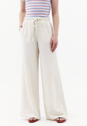Persona che indossa pantaloni beige a vita alta e gamba larga con coulisse, t-shirt a righe e sandali infradito rossi, in piedi su sfondo bianco.