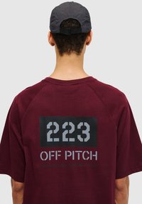 Bordeaux Baumwoll-T-Shirt mit einem großen grauen Druckdesign auf der Rückseite, das "223 OFF PITCH" und Koordinaten zeigt. Kurze Ärmel und lässige Passform.