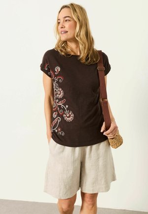 Frau trägt ein schwarzes T-Shirt mit floralem Stickmuster und beigen Shorts, hält eine gewebte Umhängetasche mit braunem Riemen.