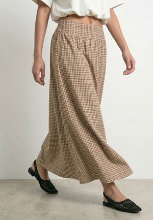 Donna che indossa pantaloni beige a quadri a gamba larga, scarpe basse nere intrecciate e un top bianco corto su uno sfondo semplice.