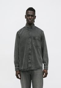 Camisa de ganga cinzenta de manga comprida com gola pontiaguda, fecho de botões à frente e bolso no peito. Textura macia e corte descontraído.