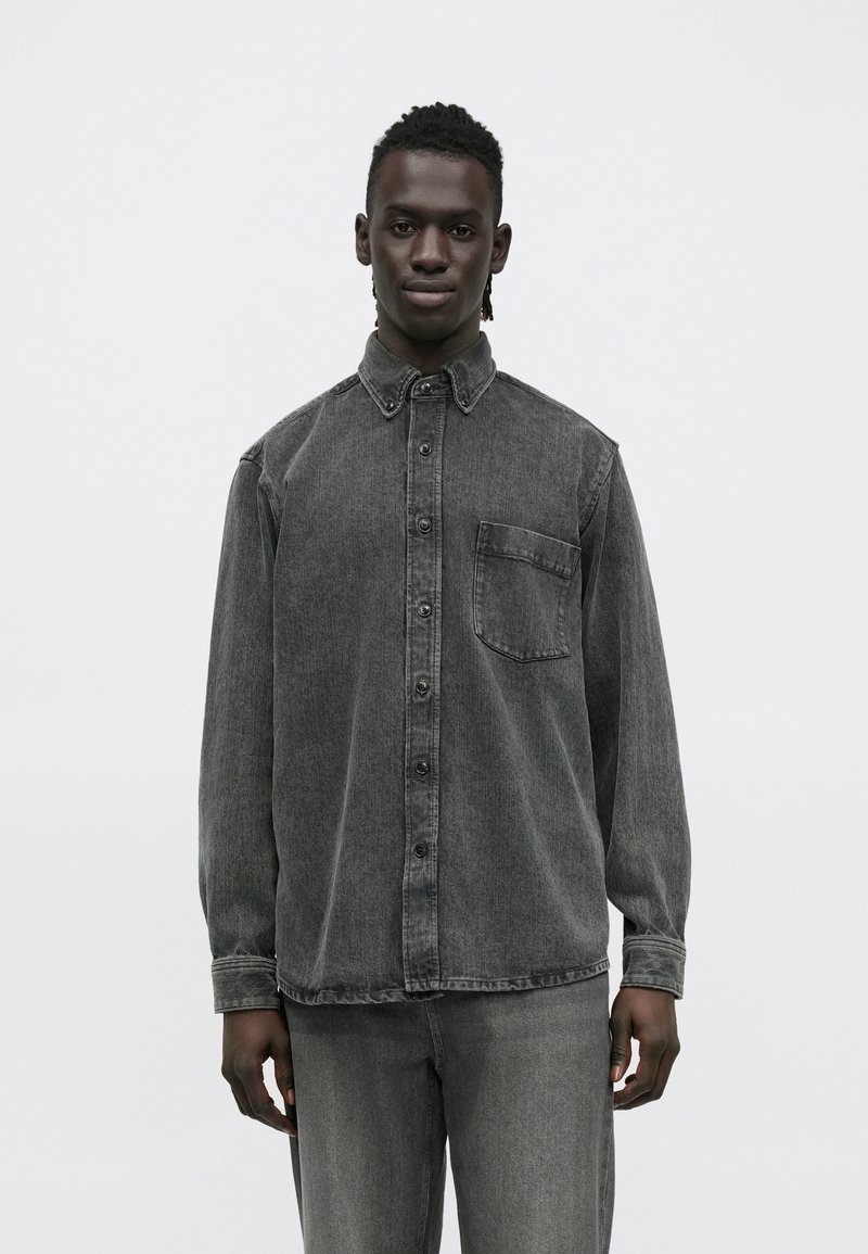 Camisa de ganga cinzenta de manga comprida com gola pontiaguda, fecho de botões à frente e bolso no peito. Textura macia e corte descontraído.