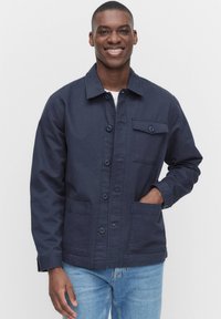 Mörkblå jacka med knäppning framtill, två framfickor och en bröstficka. Tillverkad av bomull med en slät yta.