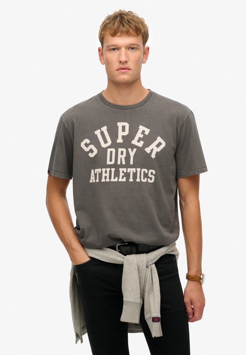 Camiseta de algodón gris con la inscripción "SUPER DRY ATHLETICS" en blanco. Presenta un cuello redondo y mangas cortas. Jeans negros y un suéter gris atado en la cintura.
