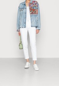 Světle modrá džínová bunda s barevnými vyšívanými vzory, bílé přiléhavé kalhoty a bílé tenisky. Malá zelená kabelka je nesena.