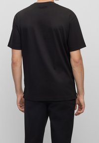 Muž v jednobarevném černém tričku s krátkým rukávem a malým logem "BOSS" na horní části zad, doplněný černými kalhotami, stojí na světlém šedém pozadí.