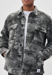 Mężczyzna w czarno-szarej, teksturowanej kurtce z guzikami, mający dwie kieszenie na piersi i ręce w kieszeniach bocznych, noszący białą koszulę.