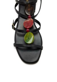 Sandalia plana negra con correas delgadas. Presenta piedras coloridas en rojo, verde y marrón a lo largo del centro. Textura suave. Punta cuadrada.