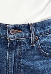 Μέση μπλε τζιν παντελονιού με ορατό κουμπί, θηλιά ζώνης και άκρη εμπρόσθιας τσέπης πάνω σε λευκό υφασμάτινο φόντο.