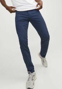 Námornícky modré slim-fit nohavice vyrobené z elastického materiálu s hladkou textúrou. Majú predné vrecká a zips na prednej strane. Nosené s sivými teniskami.