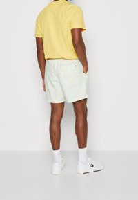 Uomo che indossa una camicia gialla a maniche corte, pantaloni corti a fantasia pastello, calzini bianchi e sneaker bianche, in piedi con le mani in tasca, di spalle.