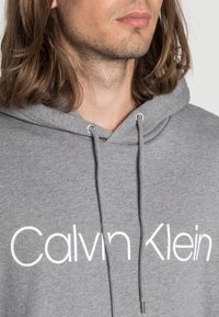 Szara bluza z kapturem ze sznurkiem, z dużym białym logo "Calvin Klein" na przodzie. Wykonana z miękkiego materiału i o standardowym kroju.