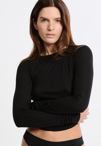 Donna con lunghi capelli castani, che indossa un top a maniche lunghe nero aderente e della biancheria intima nera, in piedi con le braccia incrociate su uno sfondo bianco.