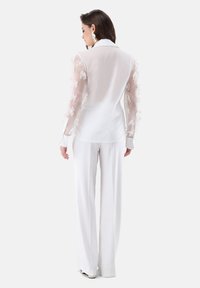 Weiße Bluse mit transparenten, bestickten Ärmeln mit floralen Motiven. Kombiniert mit weißen, weit geschnittenen Hosen und einem Kragen. Glatte Textur.