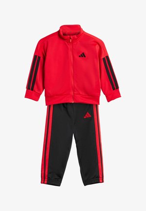 Punainen Adidas -treeniasu, jossa on vetoketjulla varustettu takki, jossa on mustat raidat hihoissa, sekä mustat housut, joissa on punaiset sivuraidat ja logo.
