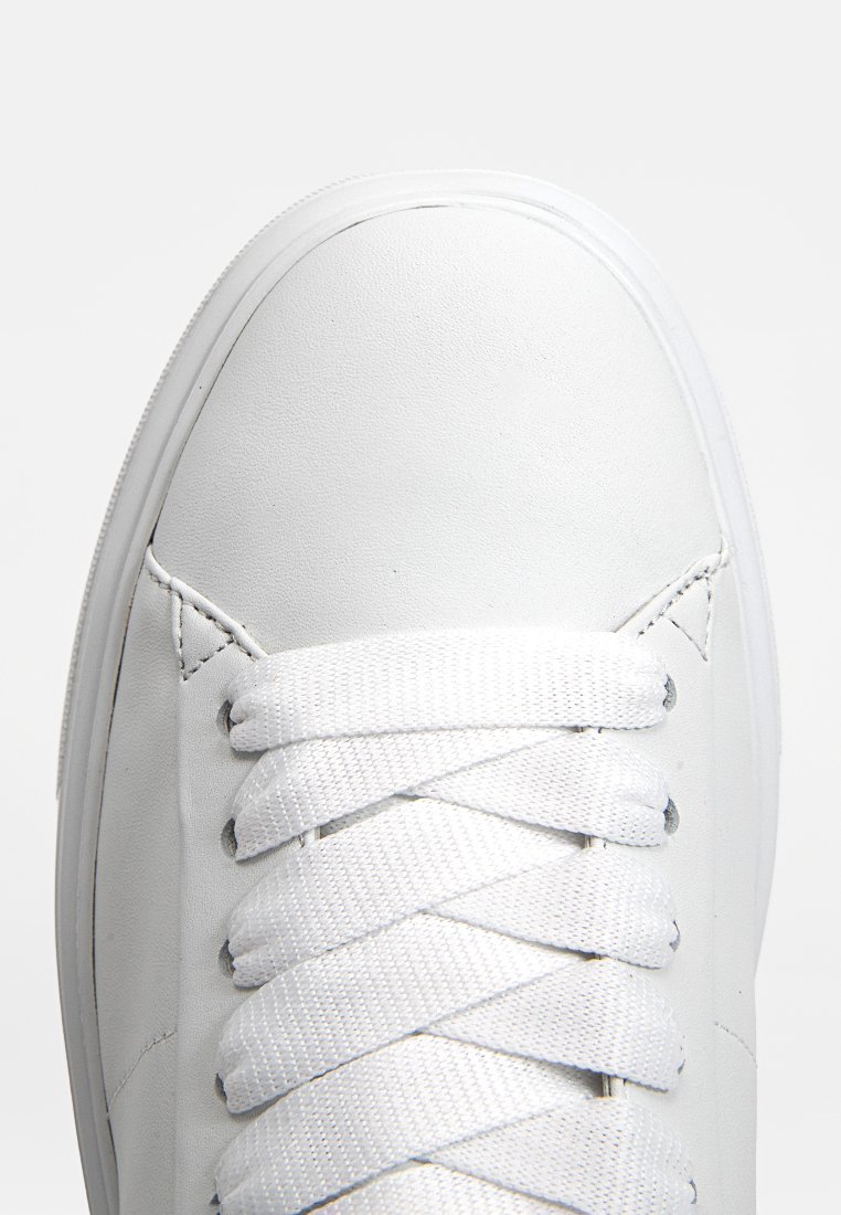 Sneaker blanc avec une tige en cuir lisse, bout rond et lacets blancs plats enfilés à travers six œillets. Aucun logo ni motif visible.