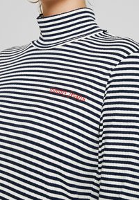 Sweter z golfem w paski z ciemnogranatowymi i białymi poziomymi paskami, żebrowaną teksturą oraz czerwoną haftowaną aplikacją "tommy jeans" na klatce piersiowej.