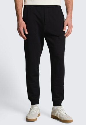 Mężczyzna w czarnych dopasowanych spodniach jogger i białych sneakersach stoi na jasnym tle.