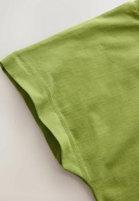 Detailní záběr zeleného bavlněného trička s hladkou látkou a zdvojeným prošitým lemem na světlém pozadí.