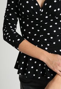 Czarna bluzka w białe kropki; ma długie rękawy, dekolt w kształcie litery V, marszczenia w okolicy biustu oraz rozkloszowany dół w stylu peplum.