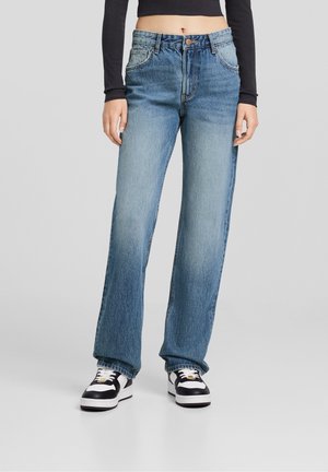Kvinde iført falmede, lige blå jeans, sort cropped langærmet top og sort-hvide sneakers, stående mod en ensfarvet baggrund.