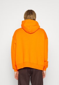 Sudadera grande naranja con capucha, hombros caídos y dobladillo acanalado. La tela parece suave y gruesa, proporcionando una cobertura acogedora.