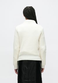 Valkoinen neulottu villapaita, jossa on kuvioitu pinta ja suora leikkaus. Pitkät hihat ja kaulus. Päällä mustan nahkahameen päällä.